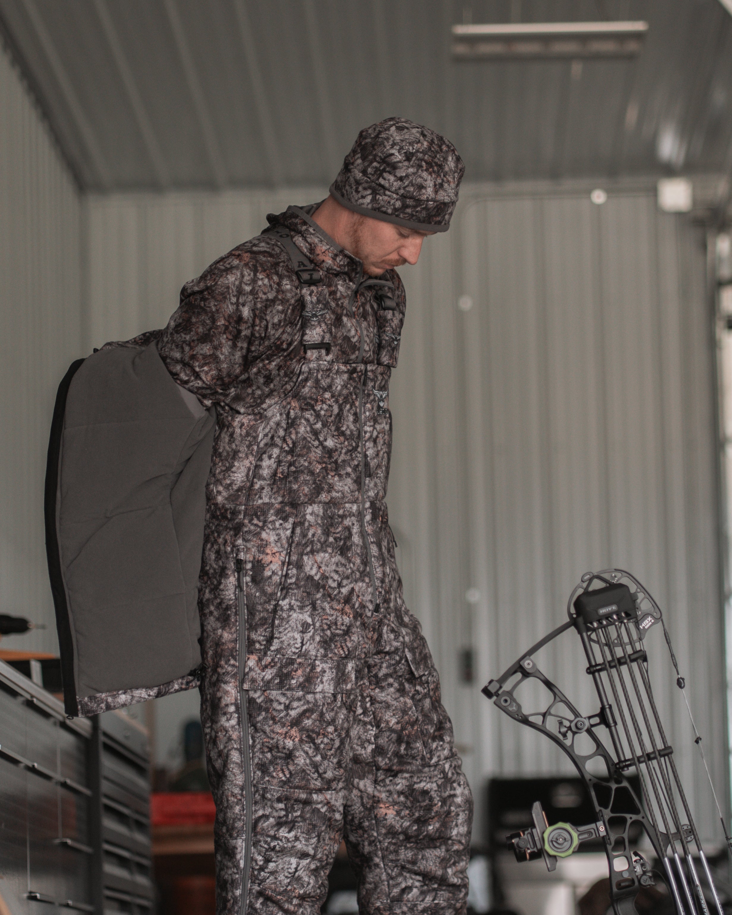 Hunter in full RAPTOR camo Heavyweight Bib from ASIO Gear layering up in gear room beside compound bow