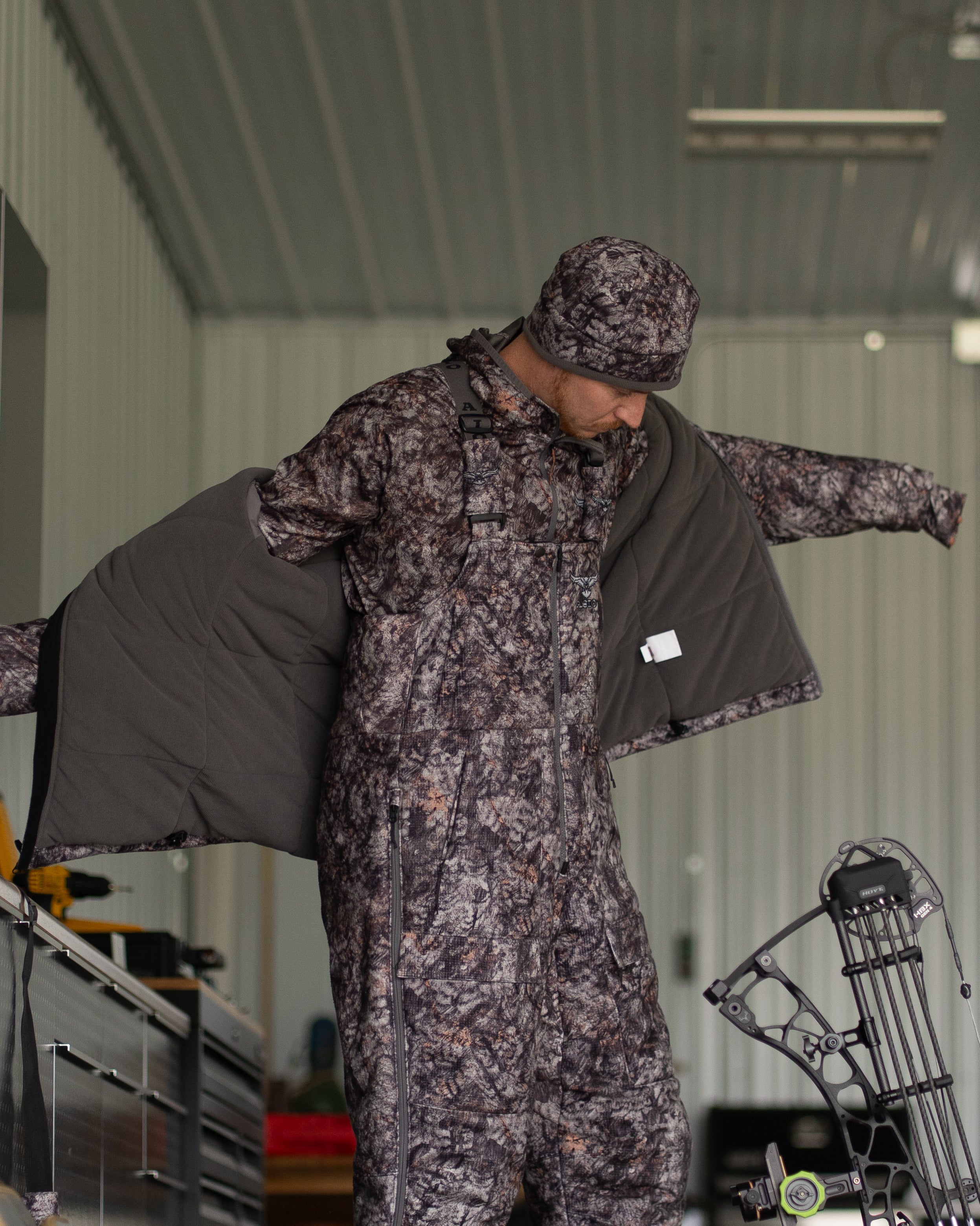 Hunter in ASIO Gear RAPTOR camo heavyweight bibs and jacket preparing gear in indoor setup with compound bow