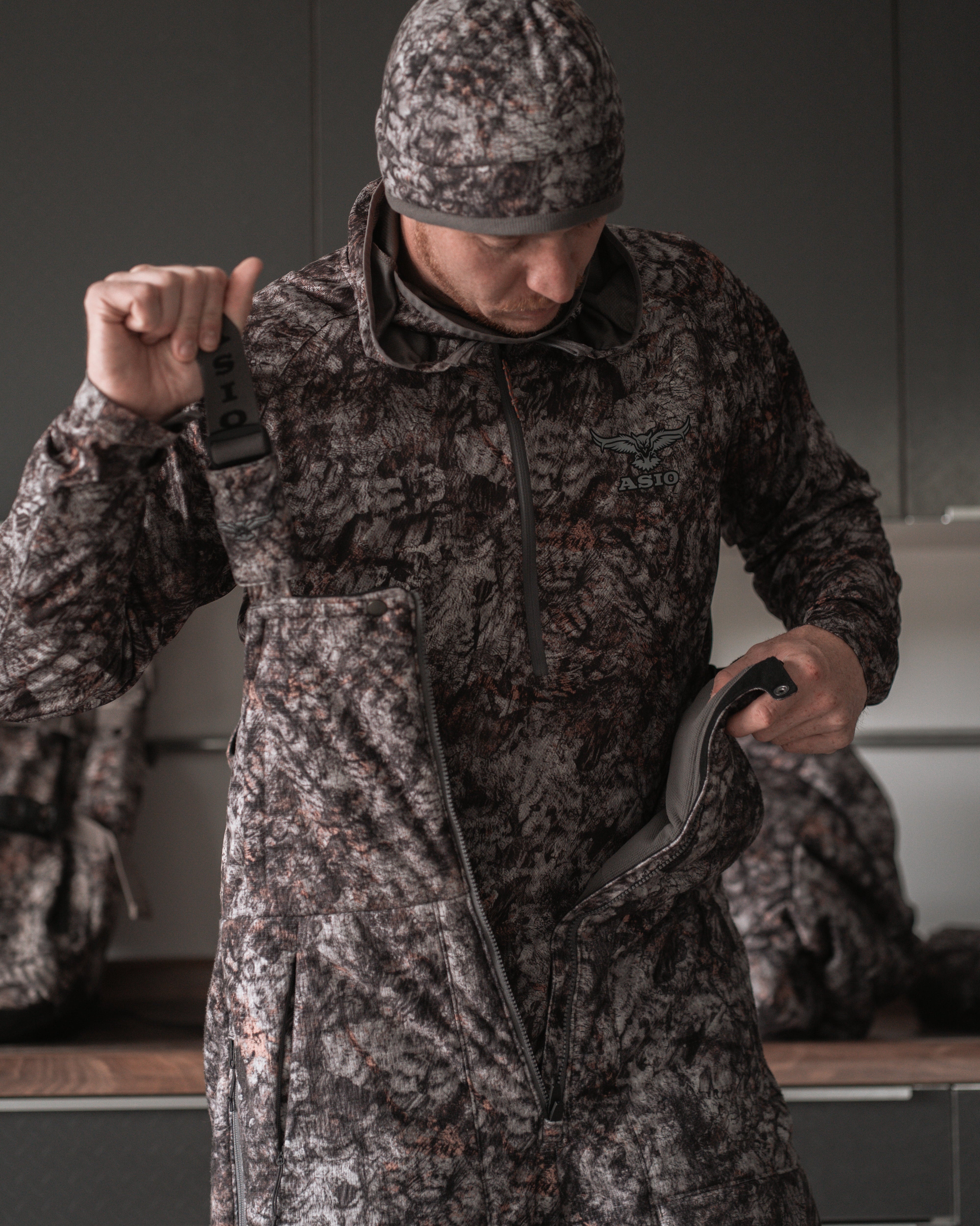 Hunter wearing ASIO Gear Heavyweight Bib in RAPTOR camo, showing front zip and branded suspenders