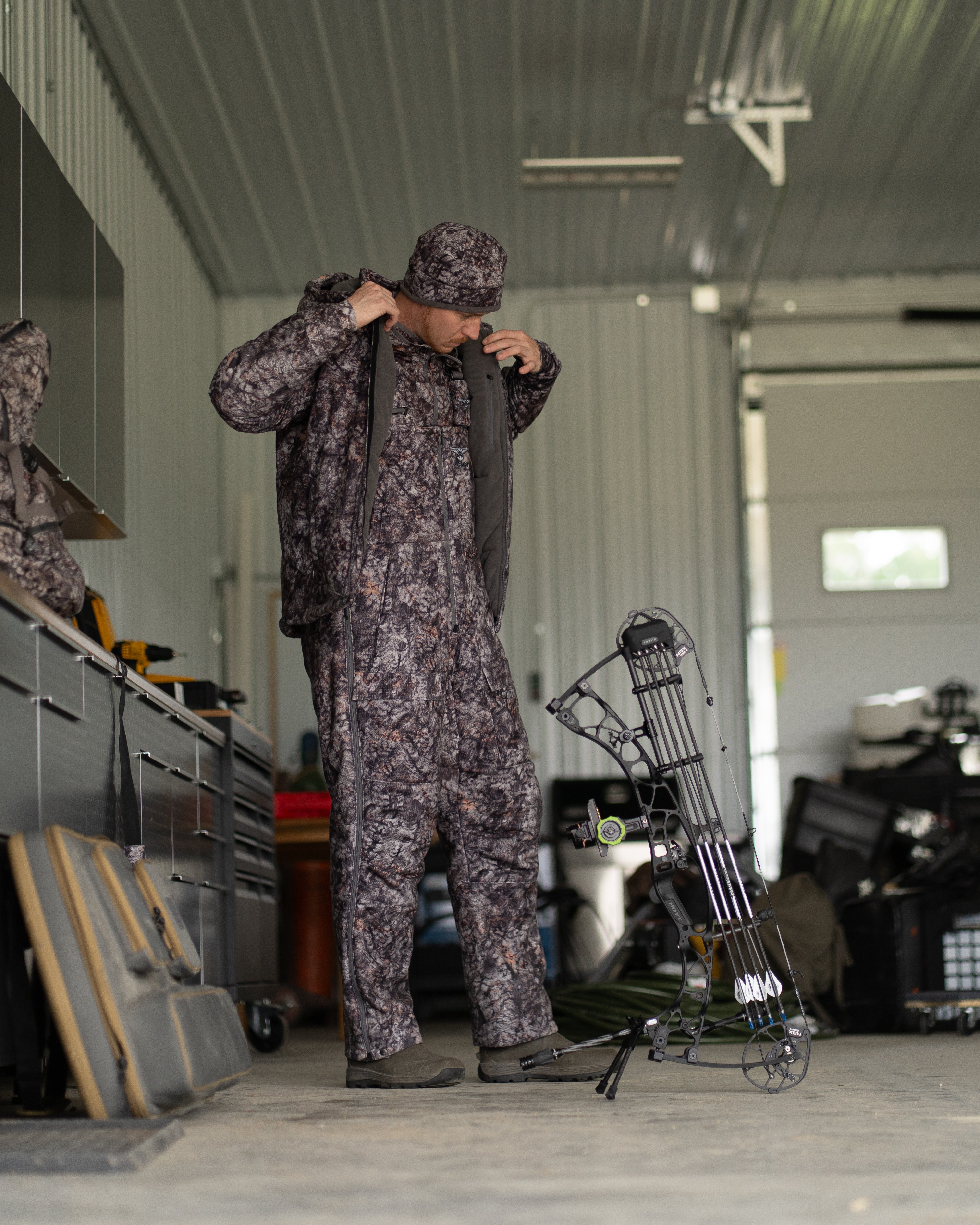 Hunter wearing ASIO Gear RAPTOR camo heavyweight bib and jacket preparing gear indoors next to compound bow
