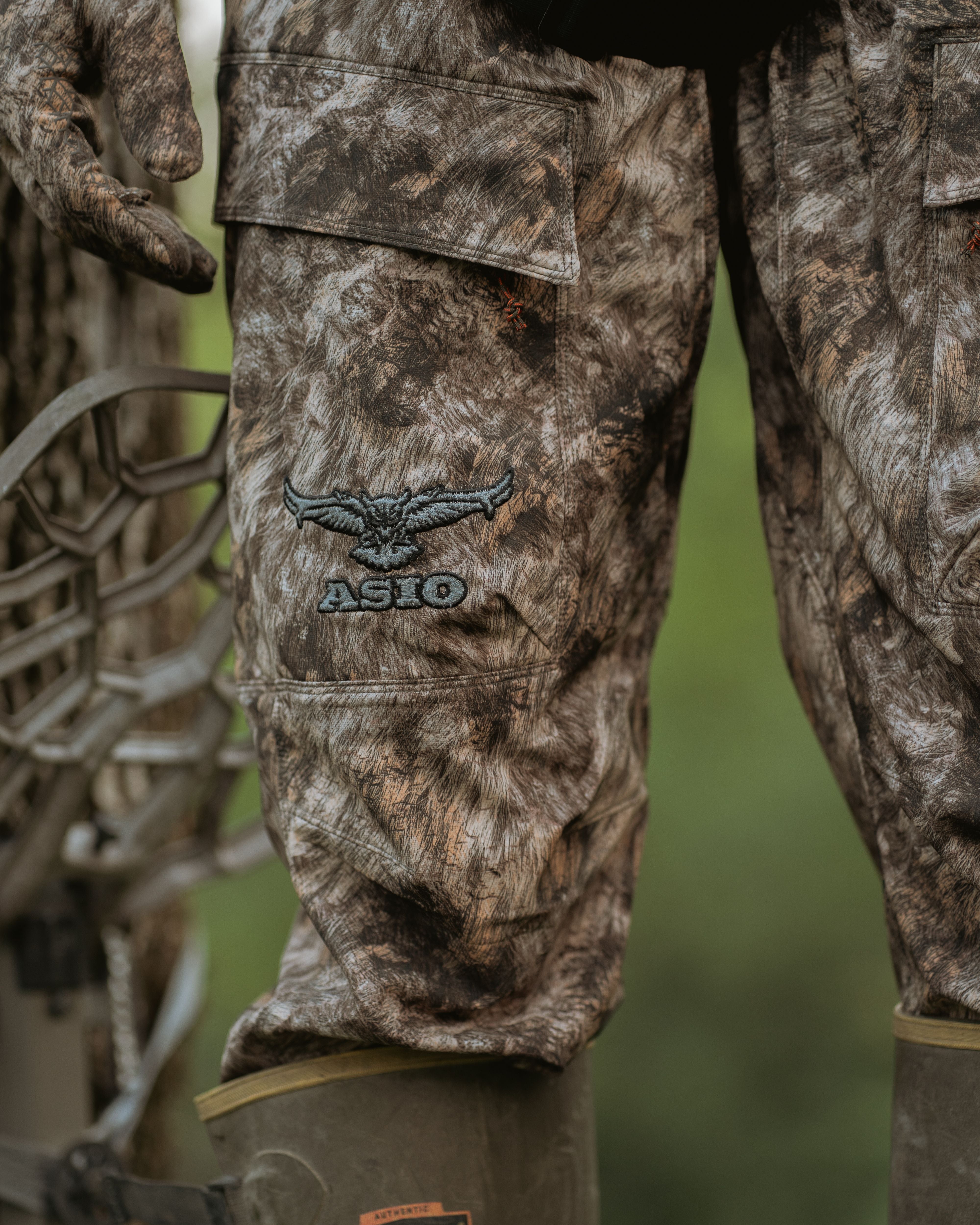 Hunter standing in treestand unzipping thigh pocket of ASIO Gear lightweight pant in RAPX camo with bow and Lacrosse boots