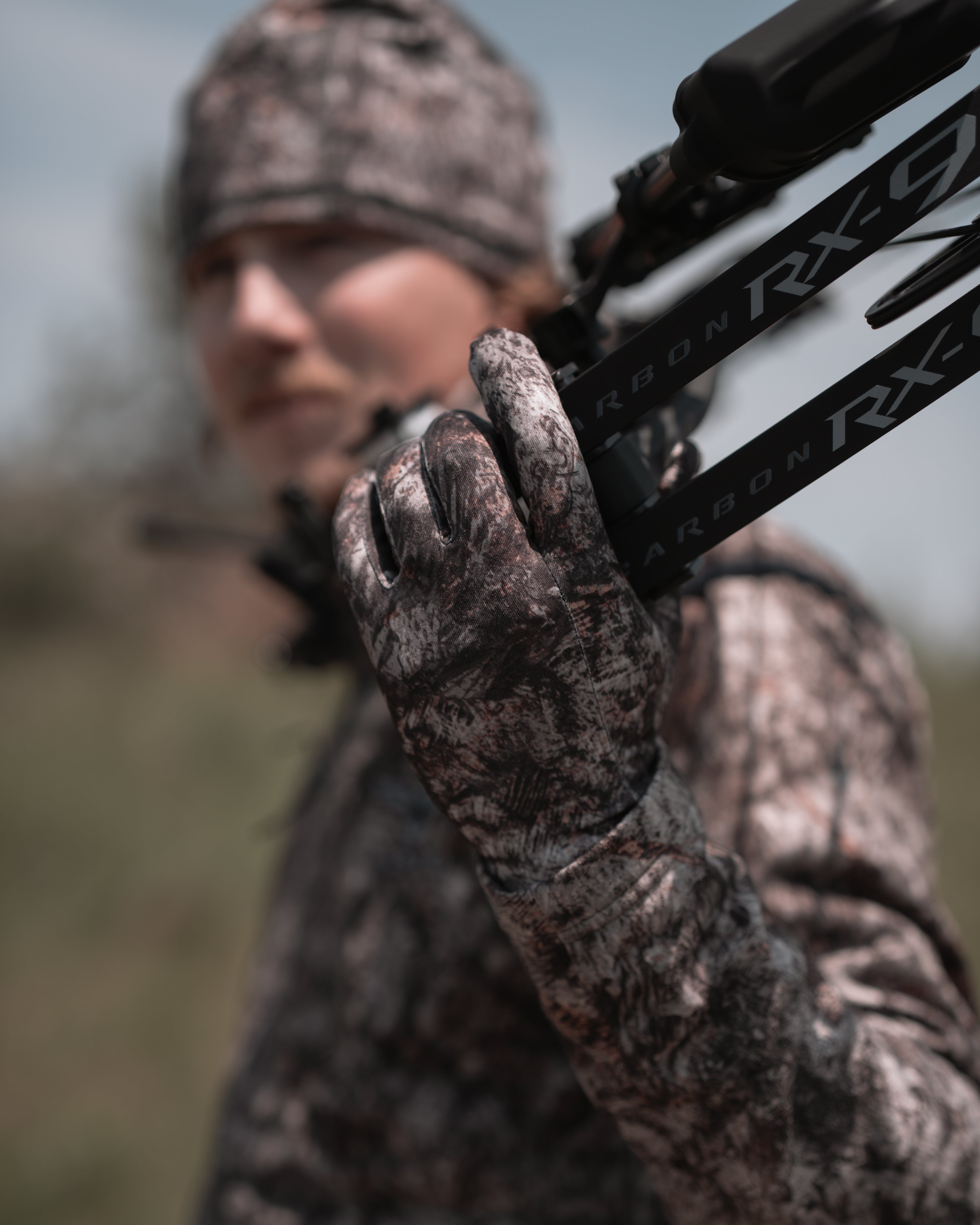 Hunter in ASIO RAPTOR camo with bow over shoulder wearing lightweight stretch gloves during active early season hunt