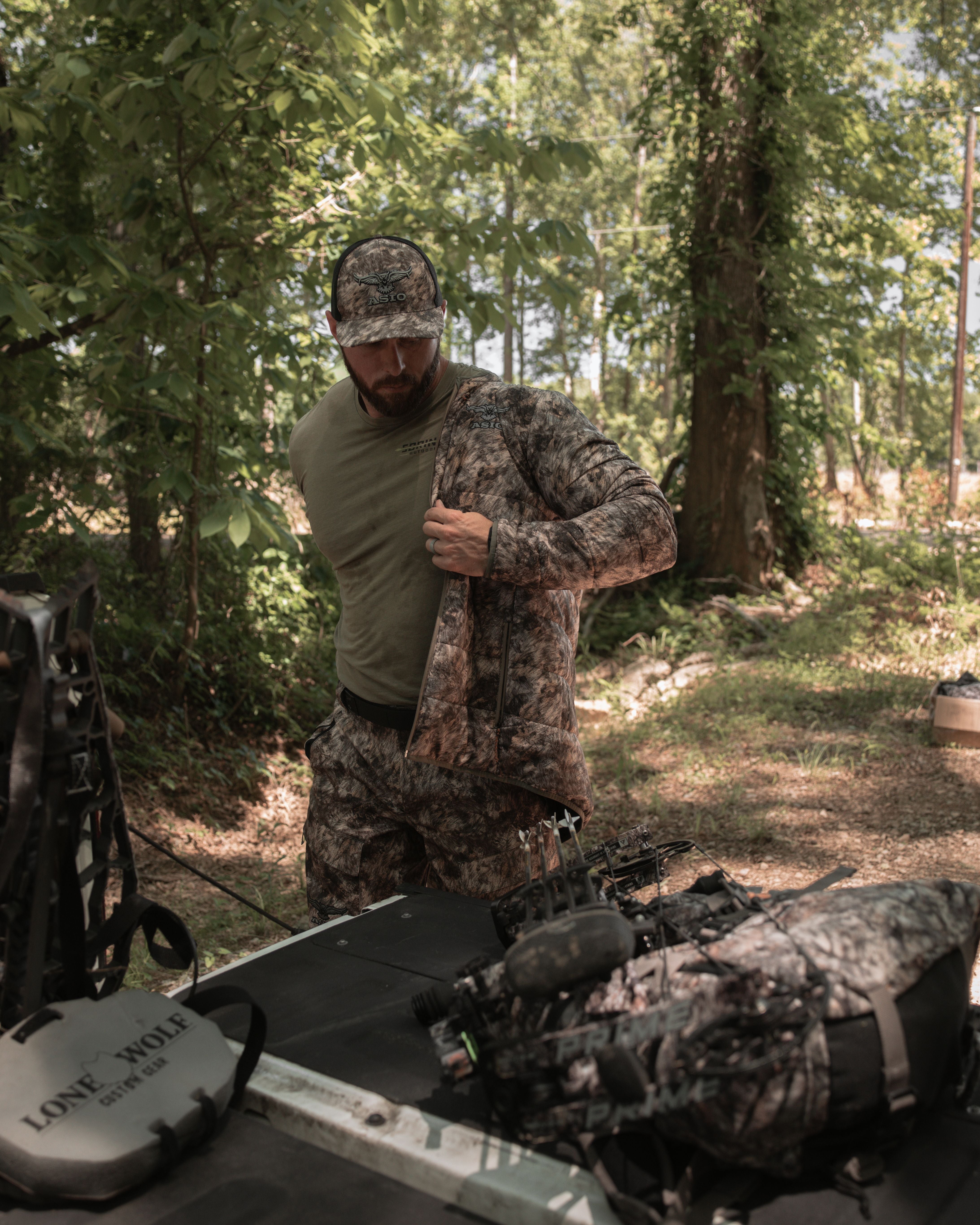 Bowhunter putting on ASIO RAPX camo midweight puffy jacket next to pack and compound bow in wooded hunting area