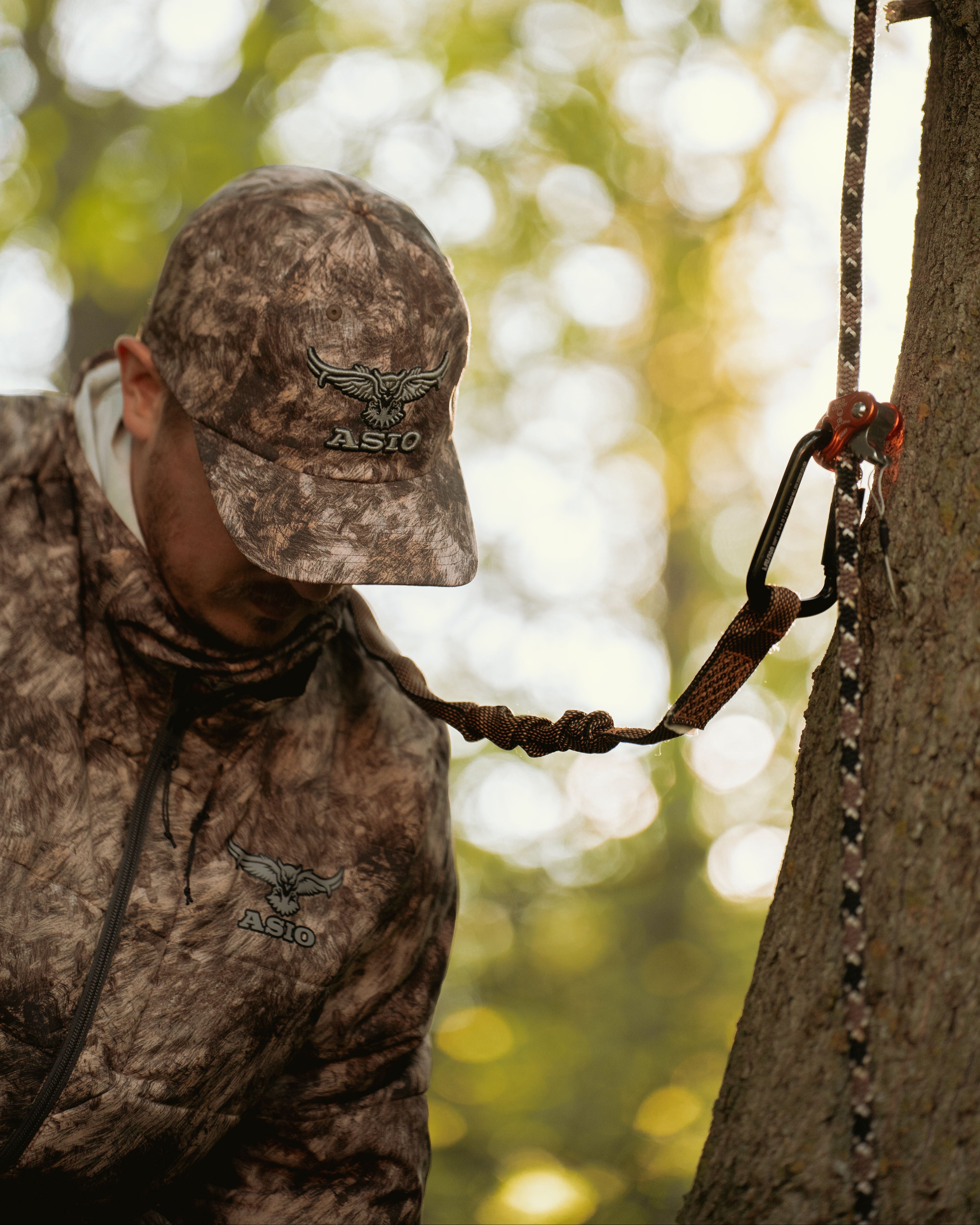 Hunter wearing ASIO RAPX camo midweight puffy jacket and cap securing treestand safety harness around tree at sunrise