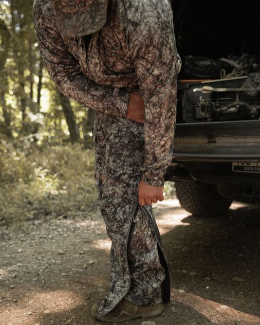 Bowhunter adjusting sherpa pant zip over boots for easy layering in ASIO Gear Midweight Sherpa Pant Raptor camo.
