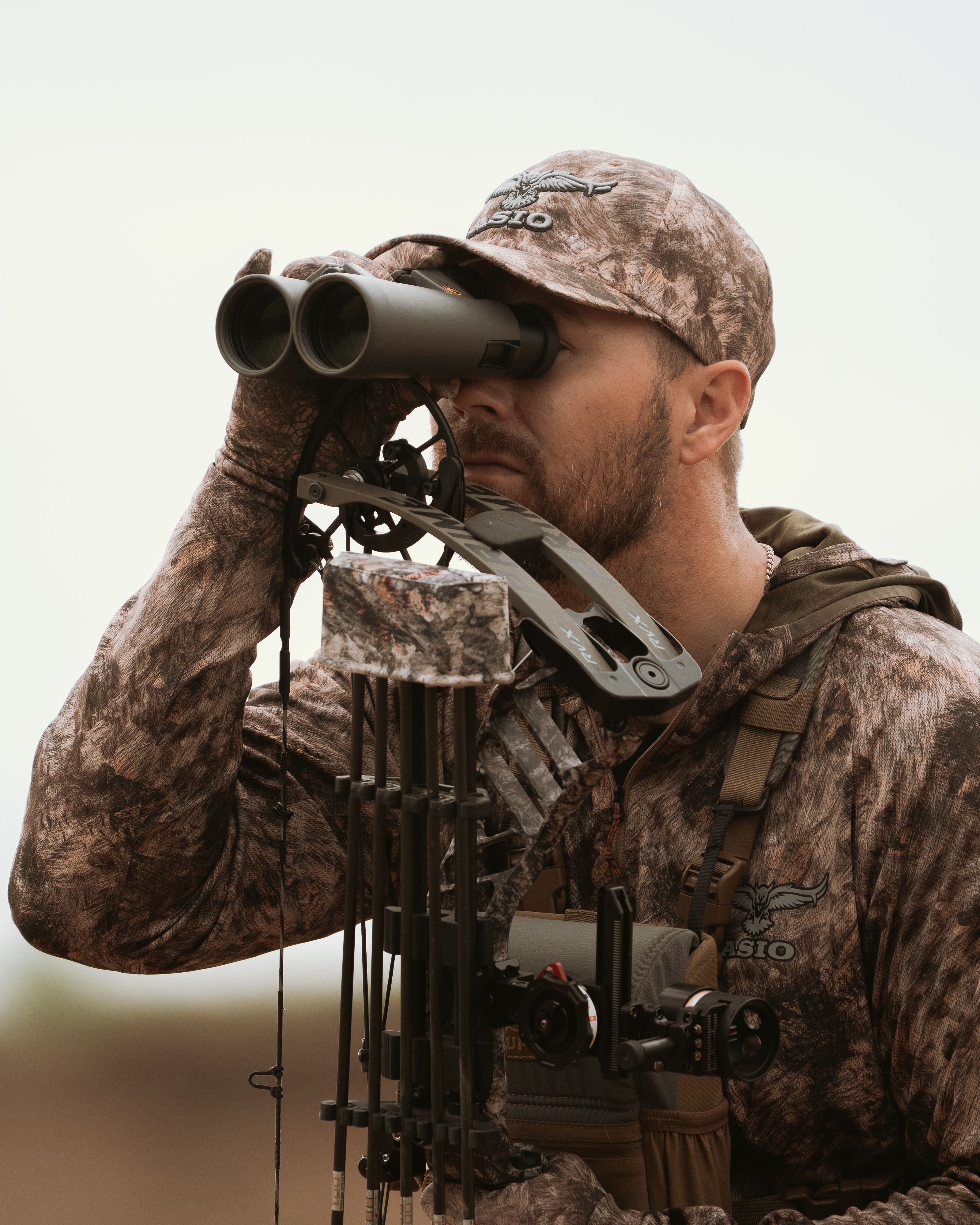  Bowhunter glassing in open terrain wearing ASIO Midweight Hoodie in RAPX Camo and matching hat.